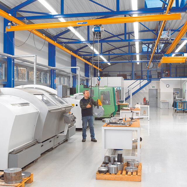 Mitarbeiter bedient ein Hängebahnsystem mit Kettenzug und bewegt eine Last in einer Werkhalle