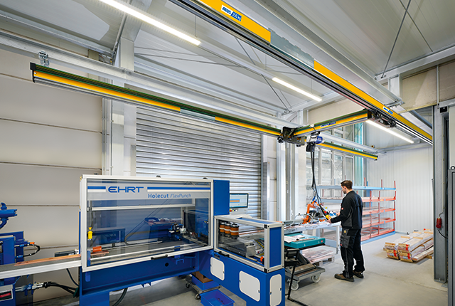 Einträgerkran EHB-X bewegt eine Last in einer Industriehalle, während ein Mitarbeiter im Hintergrund an einer Maschine arbeitet.